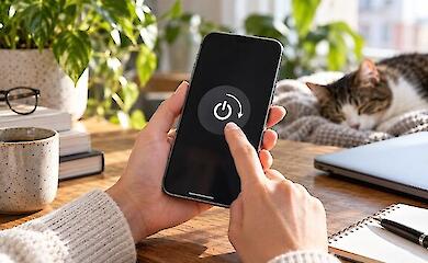 Hands hold a smartphone with a reboot symbol on the screen; nearby a laptop, a cup, and a sleeping cat