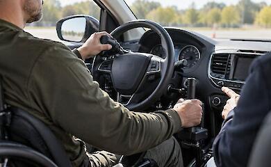 A driving-school student operates a car with adapted hand controls while an instructor sits nearby.