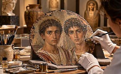 A conceptual round wooden painting of two young men on a restoration table, with brushes and a conservator&rsquo;s gloved hands nearby